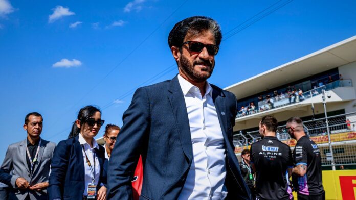 Mohammed Ben Sulayem walking confidently along the Formula 1 paddock, flanked by officials and team personnel under a clear blue sky.